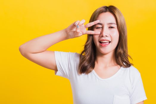 Asian Thai happy portrait beautiful cute young woman smile standing wear t-shirt showing finger making v-sign symbol near eye looking to camera, isolated studio shot yellow background with copy space