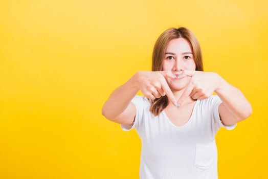 Asian Thai happy portrait beautiful cute young woman smile standing make finger heart figure symbol shape sign with two hands and looking camera, studio shot isolated yellow background with copy space