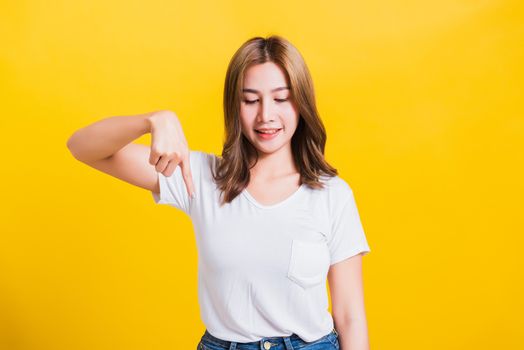 Asian Thai happy portrait beautiful cute young woman standing wear t-shirt makes gesture two fingers point below down and looking to down, studio shot isolated on yellow background with copy space