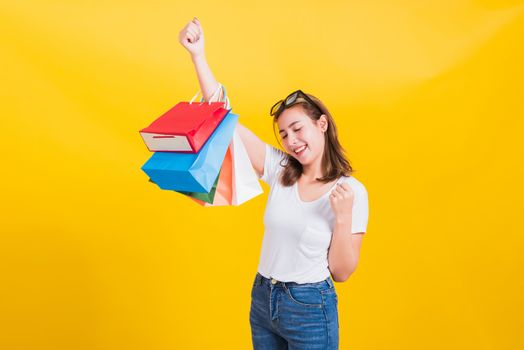 Asian Thai portrait happy beautiful cute young woman smiling stand with sunglasses excited holding shopping bags multi color looking camera, studio shot isolated yellow background with copy space