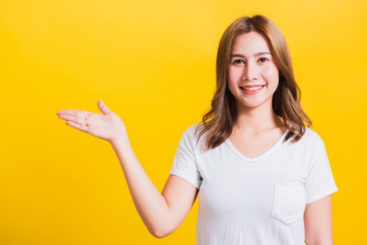 Asian Thai happy portrait beautiful cute young woman standing wear white t-shirt holding something on palm away side looking to camera, studio shot isolated on yellow background with copy space