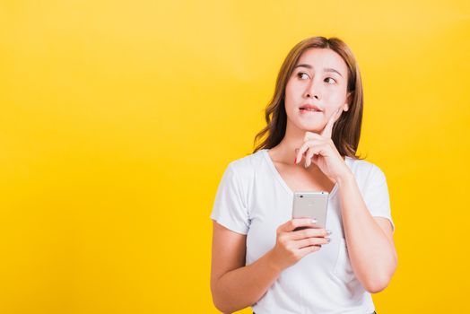 Asian Thai portrait happy beautiful cute young woman stand wear t-shirt her using holding smart mobile phone and chin handle thinking looking to side, studio shot isolated yellow background copy space