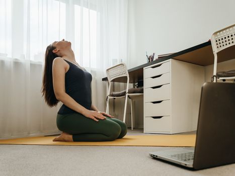 Young woman sitting on floor and doing yoga or stretching at home near laptop. Yoga online in small space home and wellbeing concept. Copy space for text or design.