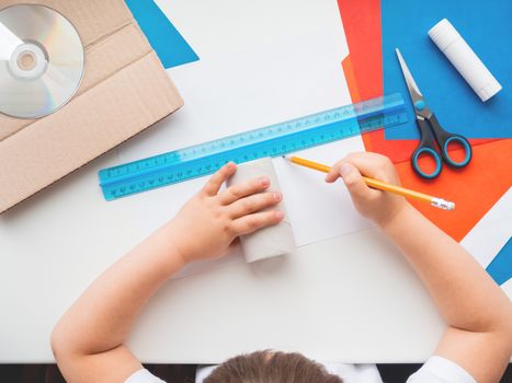 Child makes something out of colored paper, toilet paper core, carton, scissors, glue, CD-disk, pencil and ruler. Red, white and blue pallette. Making toys from improvised materials.