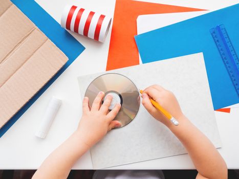 Child makes something out of colored paper, toilet paper core, carton, scissors, glue, CD-disk, pencil and ruler. Red, white and blue pallette. Making toys from improvised materials.