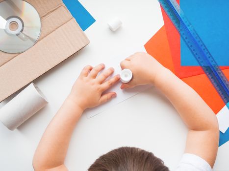 Child makes something out of colored paper, toilet paper core, carton, scissors, glue, CD-disk, pencil and ruler. Red, white and blue pallette. Making toys from improvised materials.