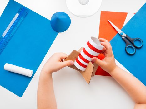 Child makes colorful rocket out of colored paper and toilet paper carton core. Also he needs scissors, glue, CD-disk, pencil and ruler. Red, white and blue handmade spaceship. Making toys from improvised materials.