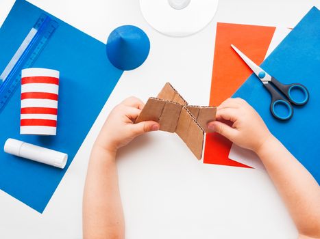 Child makes colorful rocket out of colored paper. Also he needs carton, scissors, glue, CD-disk, pencil and ruler. Red, white and blue handmade spaceship. Making toys from improvised materials.