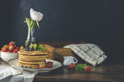 Pancake with srtawberry, blueberry and mint in ceramic dish, syrup from small ceramic jar and flowers on a dark wooden table and black background. 