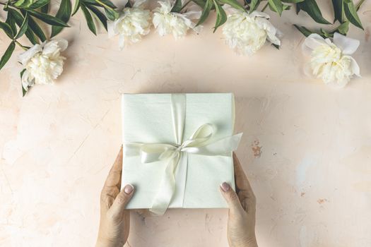 Womans hands hold gift on light concrete table surface surrounded beautiful white peony flowers. Top view, flat lay, greeting card. Feminine flat lay composition with flowers and present.