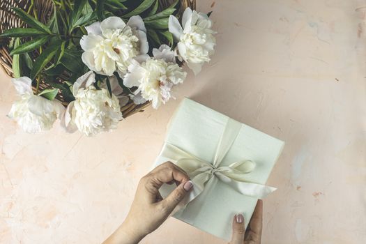 Woman opening her present, top view. Female's hands pull ribbon to unwrap gift box among the white peony flowers, festive flat lay arrangement.