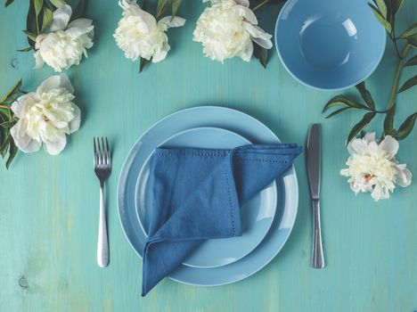 Flat lay composition with white peony flowers, empty plate with napkin and cutlery on wooden turquoise table surface, festive flat lay arrangement.