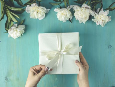 Woman opening her present, top view. Female's hands pull ribbon to unwrap gift box among the white peony flowers on wooden turquoise table surface, festive flat lay arrangement.