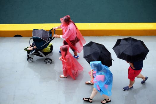 Top view of family walking under rain, covered with umbrellas, rain jackets and stroller roof.