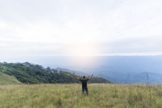 Happy young man standing with raised arms in green field on mountains