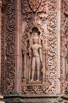 A guardian sculpture on the outer wall of a prasat, or chapel, at Banteay Srei Temple, Angkor, Cambodia. Intricate Khmer carving in red sandstone over 1000 years old.
