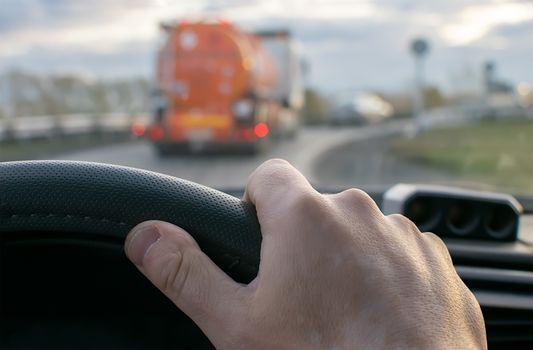 view of the driver hand at the wheel of a car against the background of a fuel tanker driving in front on the road
