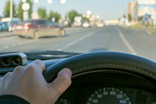 view of the driver hand on the steering wheel of a car against the background of a city road and a car leaving the secondary road on the main road