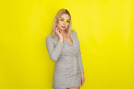 Attractive blonde woman in plaid jacket dress over yellow background wearing yellow glasses