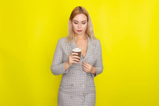 Business blonde woman wearing plaid jacket style dress and drinking coffee over yellow background