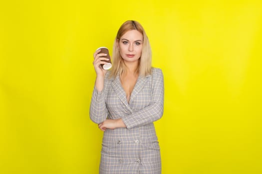 Business blonde woman wearing plaid jacket style dress and drinking coffee over yellow background