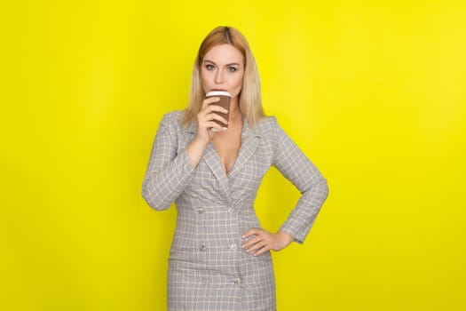 Business blonde woman wearing plaid jacket style dress and drinking coffee over yellow background