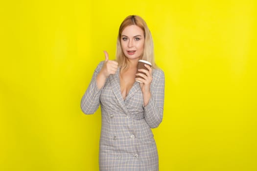 Business blonde woman wearing plaid jacket style dress and drinking coffee over yellow background