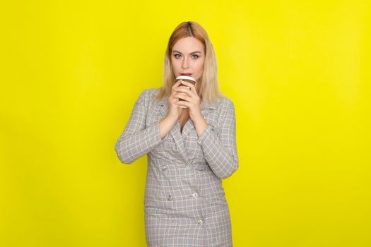 Business blonde woman wearing plaid jacket style dress and drinking coffee over yellow background