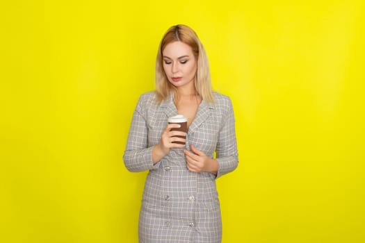 Business blonde woman wearing plaid jacket style dress and drinking coffee over yellow background