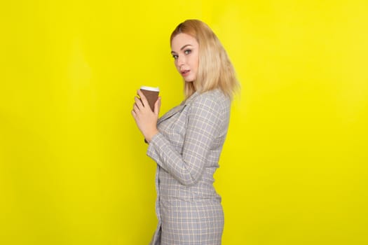 Business blonde woman wearing plaid jacket style dress and drinking coffee over yellow background