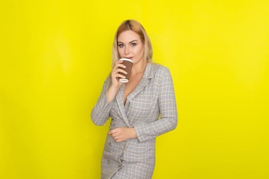 Business blonde woman wearing plaid jacket style dress and drinking coffee over yellow background