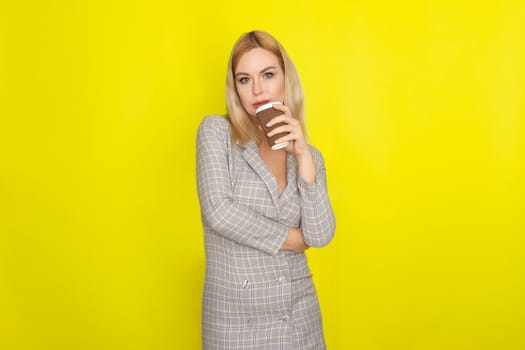 Business blonde woman wearing plaid jacket style dress and drinking coffee over yellow background