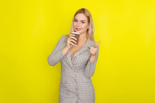 Business blonde woman wearing plaid jacket style dress and drinking coffee over yellow background