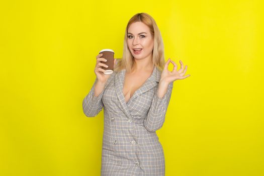 Business blonde woman wearing plaid jacket style dress and drinking coffee over yellow background