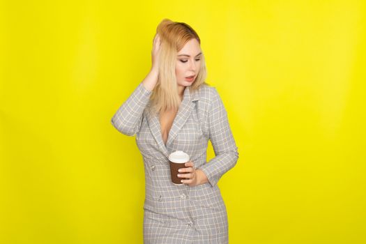 Business blonde woman wearing plaid jacket style dress and drinking coffee over yellow background