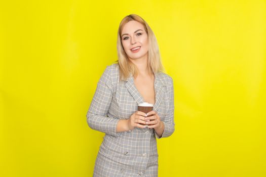 Business blonde woman wearing plaid jacket style dress and drinking coffee over yellow background