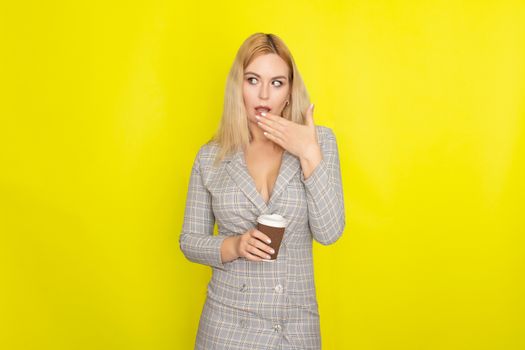 Business blonde woman wearing plaid jacket style dress and drinking coffee over yellow background