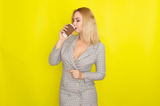 Business blonde woman wearing plaid jacket style dress and drinking coffee over yellow background