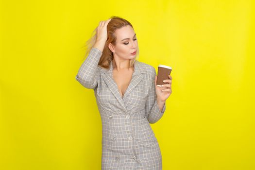 Business blonde woman wearing plaid jacket style dress and drinking coffee over yellow background