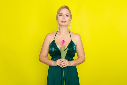 Blonde woman in green evening dress holding one tulip in her hands over yellow background