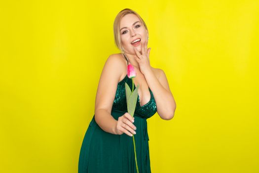 Blonde woman in green evening dress holding one tulip in her hands over yellow background