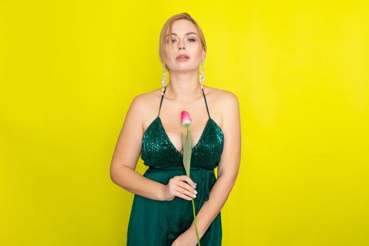 Blonde woman in green evening dress holding one tulip in her hands over yellow background