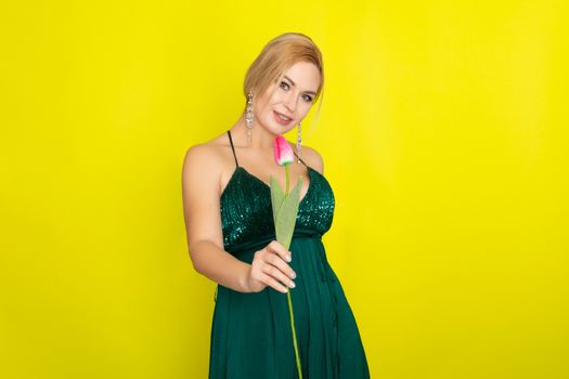 Blonde woman in green evening dress holding one tulip in her hands over yellow background