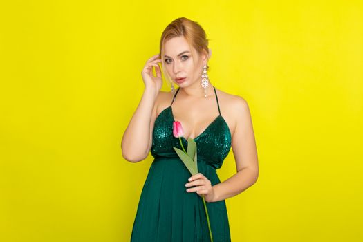 Blonde woman in green evening dress holding one tulip in her hands over yellow background