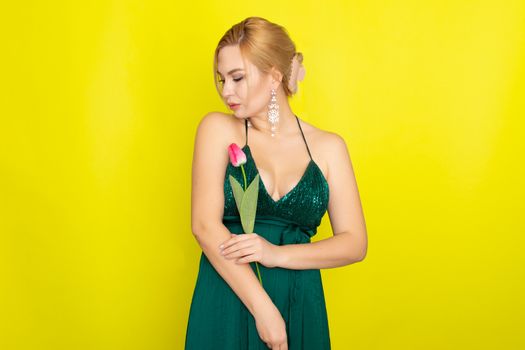 Blonde woman in green evening dress holding one tulip in her hands over yellow background