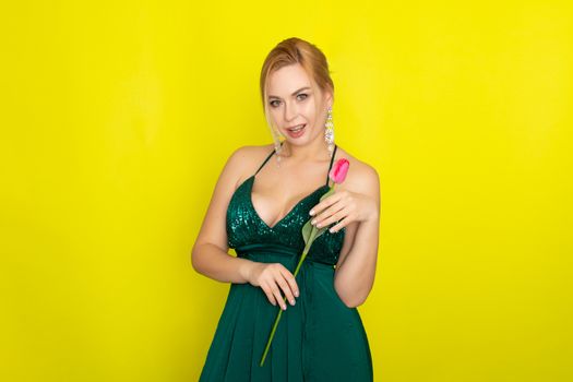 Blonde woman in green evening dress holding one tulip in her hands over yellow background