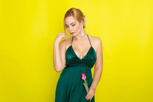 Blonde woman in green evening dress holding one tulip in her hands over yellow background