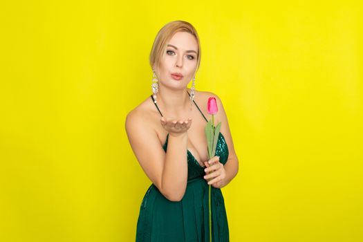 Blonde woman in green evening dress holding one tulip in her hands over yellow background