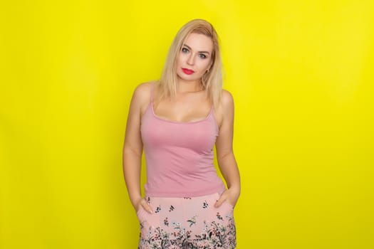 Indoor summer closeup portrait of young stylish fashion glamorous blonde woman posing in pink shorts and shirt, standing over yellow background