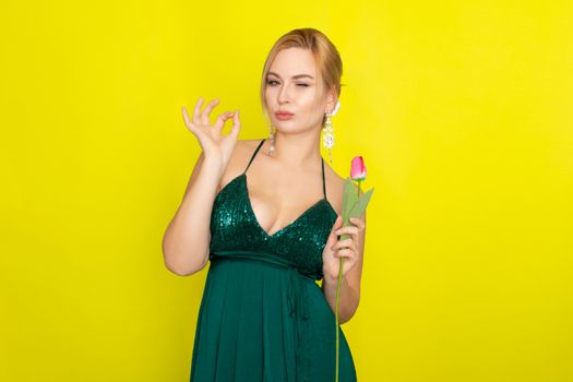 Blonde woman in green evening dress holding one tulip in her hands over yellow background